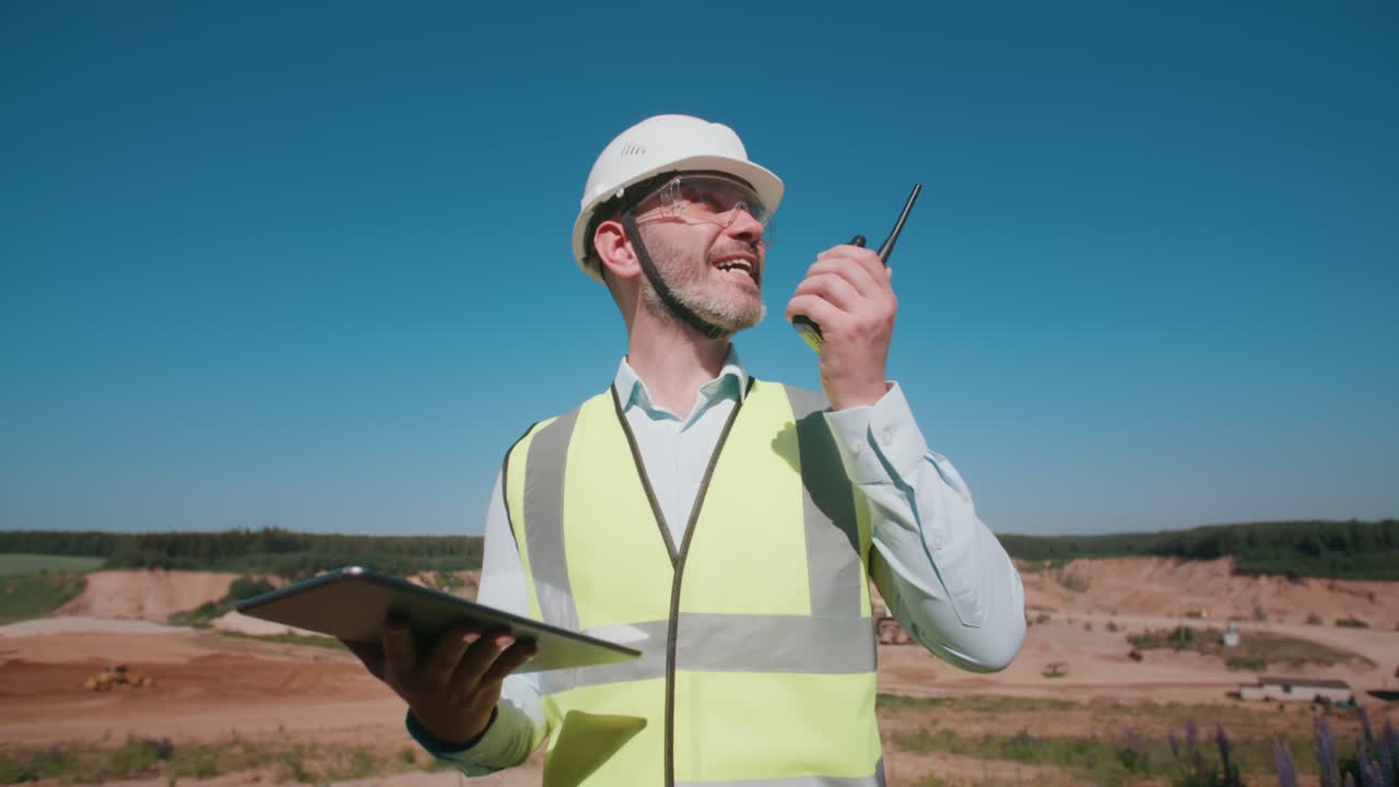 retrato de mano buen hombre trabajador de una cantera de arena, caucásico capataz barbudo instruyendo a los trabajadores usando un radio walkie talkie sosteniendo una tableta en la mano. especialista en proyectos de minería de arena en un día cálido y soleado