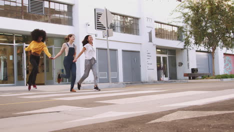 three millennial girlfriends holding hands and laughing as they run across a pedestrian crossing