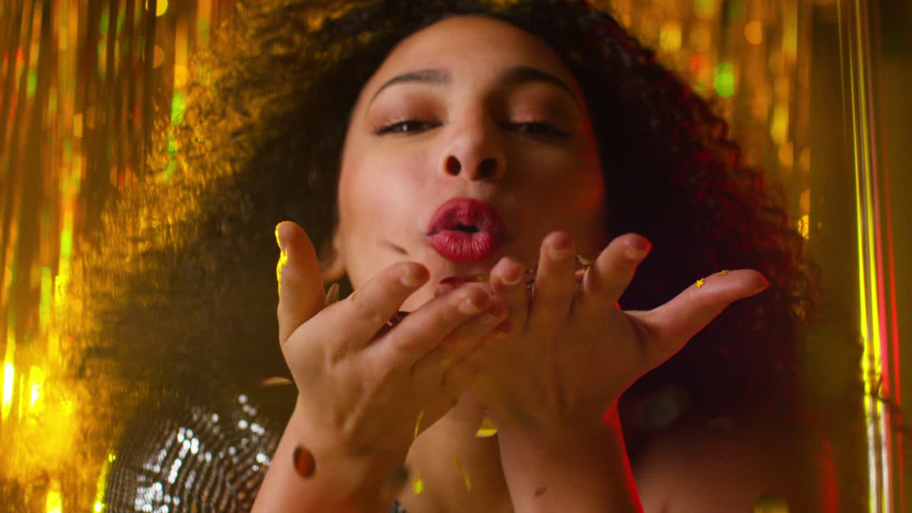 mujer joven celebrando en una fiesta o club soplando un puñado de brillo dorado hacia la cámara