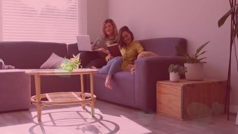 Animation-of-hearts-over-happy-caucasian-lesbian-couple-sitting-on-sofa-at-home