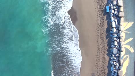 Birds-eye-view-of-waves-crashing-on-beautiful-sandy-beach-with-a-line-of-rocks-and-people-walking-also-people-cycling