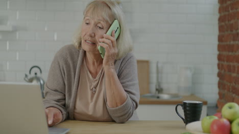 senior woman talking on phone and using laptop in kitchen