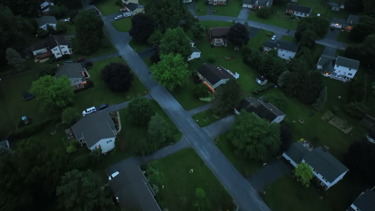 Aerial flyover housing area in idyllic green suburbia. Top down. Dusk scene in summer. One family houses and yards in quiet area of USA