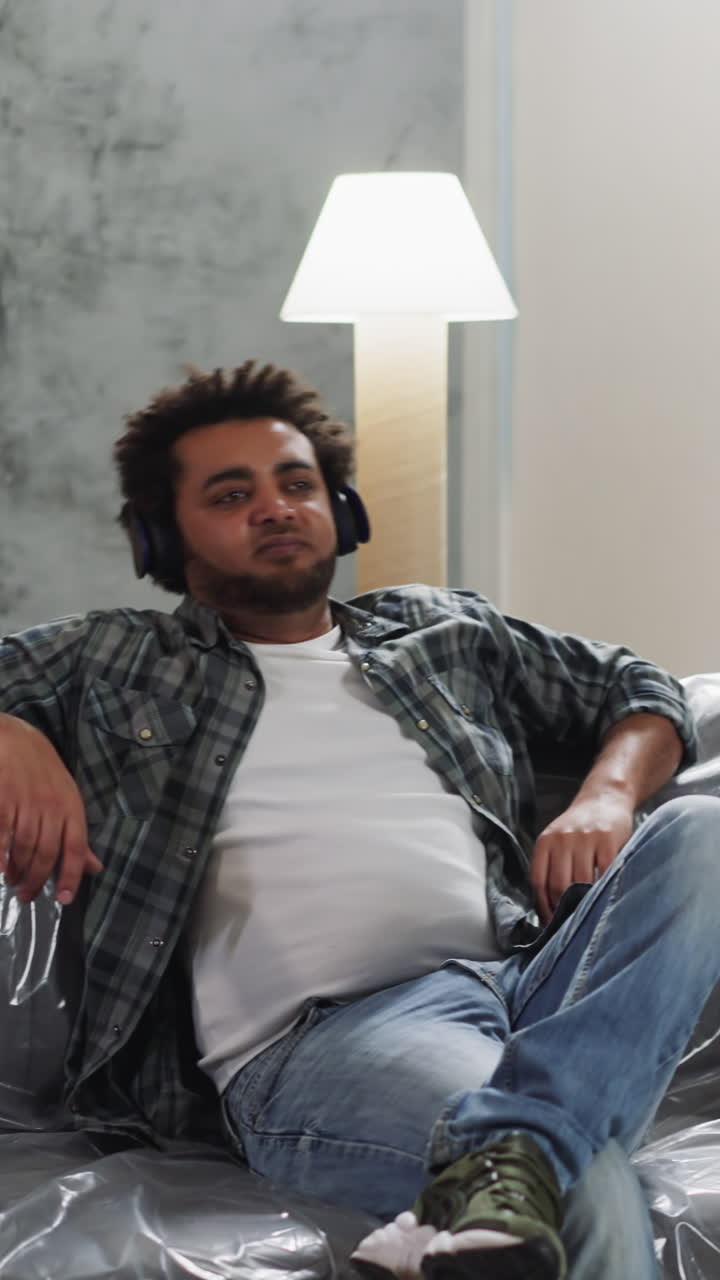 African-American repairman with earphones claps on knee sitting on foiled couch. Black man builder has break during restoration work in apartment