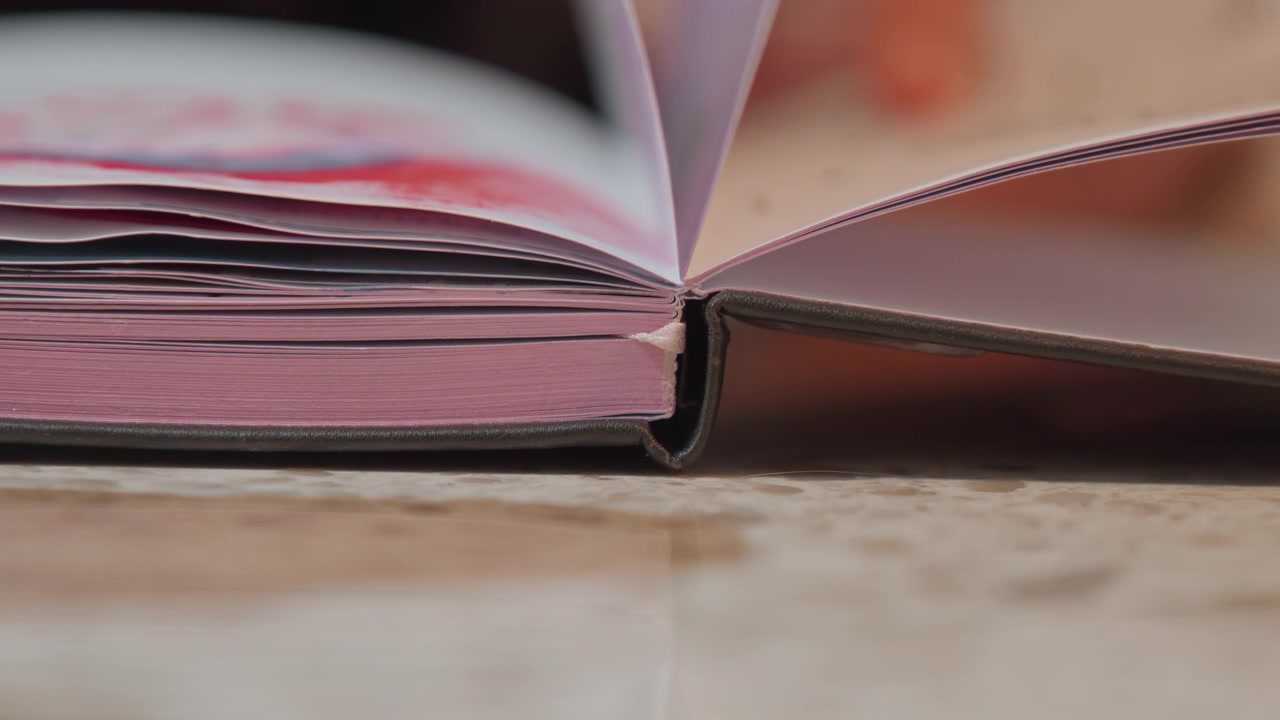 Primo piano di un libro rosa spesso con bordi delle pagine e rilegatura visibili su una superficie di colore chiaro. Lo sfondo mostra mani sfocate che suggeriscono un'attività di lettura o studio. Illuminazione soffusa.