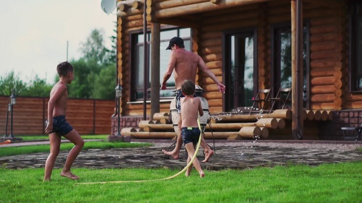 Happy family with two kids playing with garden hose pouring water on grass
