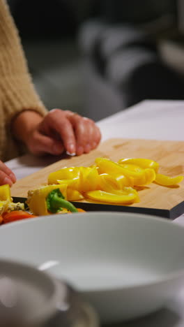 vertical video close up of woman at home in kitchen preparing healthy fresh vegetables for vegetarian or vegan meal putting sliced yellow peppers into bowl