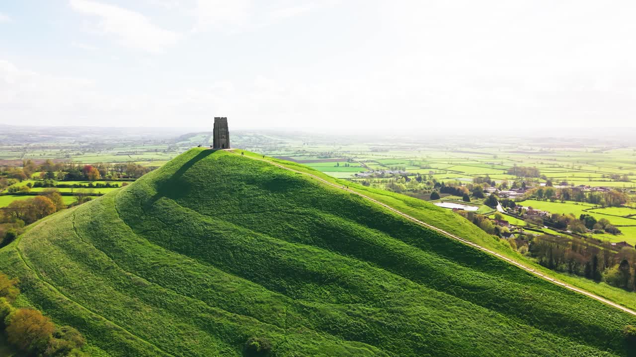 Luftfoto fra drone af Glastonbury Tor, der rejser sig over det bakkede grønne landskab i Somerset, England, med vidtstrakte landbrugsarealer, der strækker sig ud i det fjerne under blødt dagslys.
