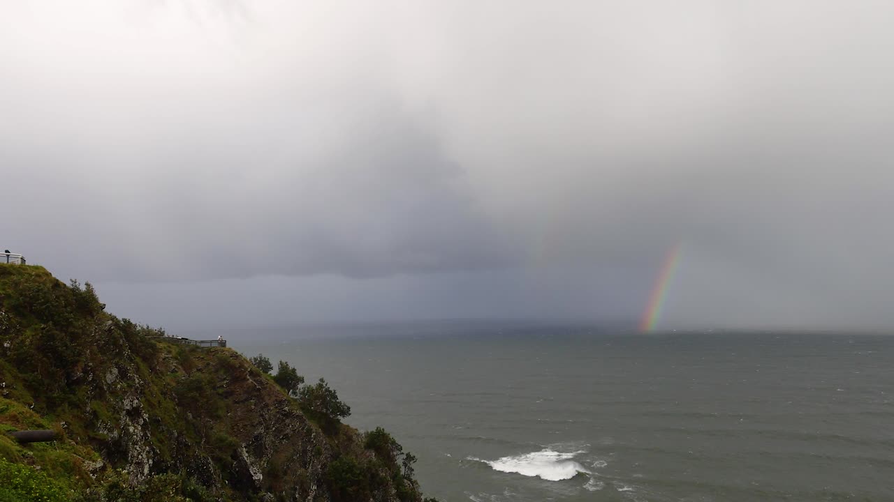 A lighthouse stands on a cliff at Byron Bay, overlooking the ocean with a faint rainbow under cloudy skies