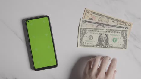 overhead currency shot of person tapping fingers next to dollar bills and green screen mobile phone