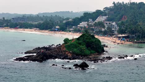 tropical sri lanka slightly elevated view of a rocky, green islet near a sandy beach and coastal town under an overcast but bright sky, captured with a standard lens