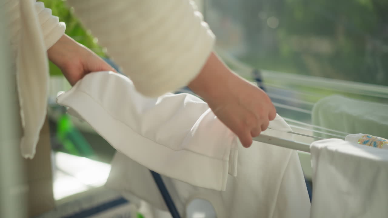 여성은 발코니에 옷 건조기에 옷을 매달고, 여성은 현대적인 평평한 느린 동작으로 빛 발코니에서 건조판에 매달린 옷을 는다.