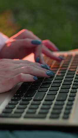 primer plano de una mujer con una camiseta rosa con uñas bien pulidas escribiendo en una computadora portátil, sombras de su mano visibles en el teclado, con un cálido resplandor de luz solar en el fondo, efecto de luz bokeh en la distancia