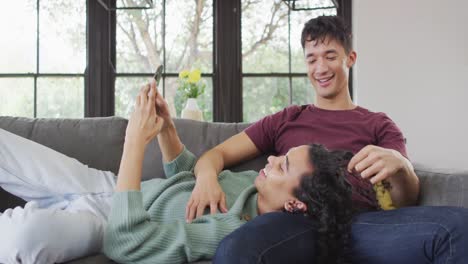 happy diverse male couple sitting on sofa, embracing and using smartphone in living room