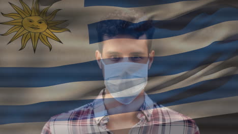 animation of flag of uruguay waving over man wearing face mask during covid 19 pandemic