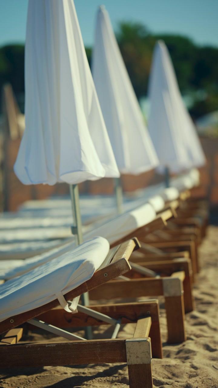 Lettini da spiaggia vuoti disposti su sabbia soffice sotto ombrelloni bianchi.
