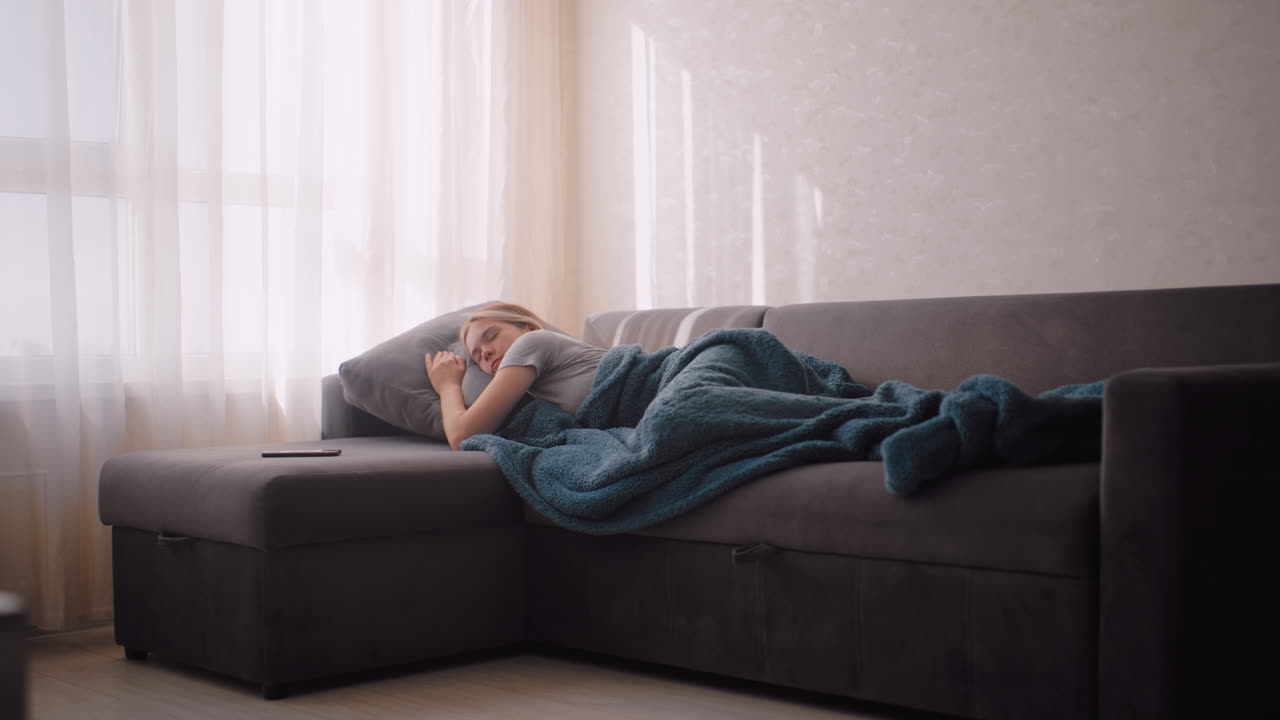 Tired woman resting on couch adjusting for comfort with pillow and blanket phone resting close by as sunlight shines through curtains creating calm serene peaceful atmosphere of relaxation