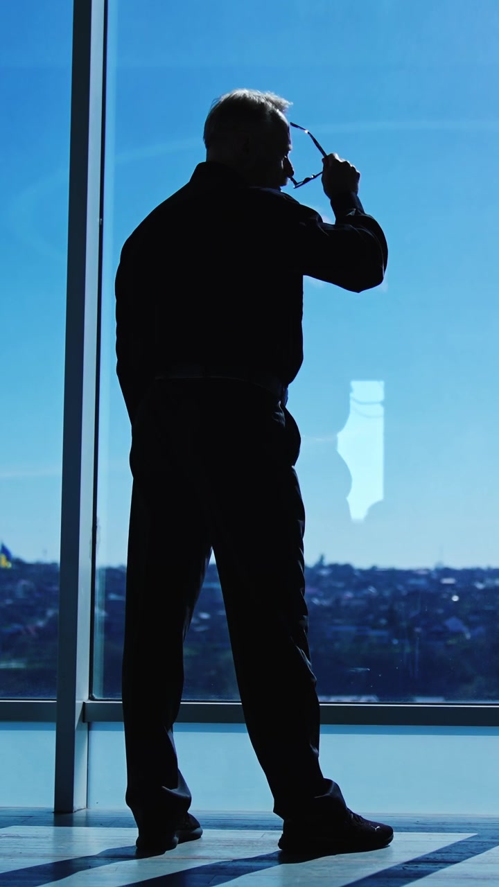 Sagoma scura di un uomo in piedi alla finestra panoramica in ufficio in una giornata di sole. Uomo d'affari che riflette su questioni, si toglie gli occhiali e guarda il paesaggio urbano. Vista dal basso. Video verticale.