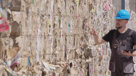 caucasian worker with tablet and blue hard hat counts stacked waste paper bales