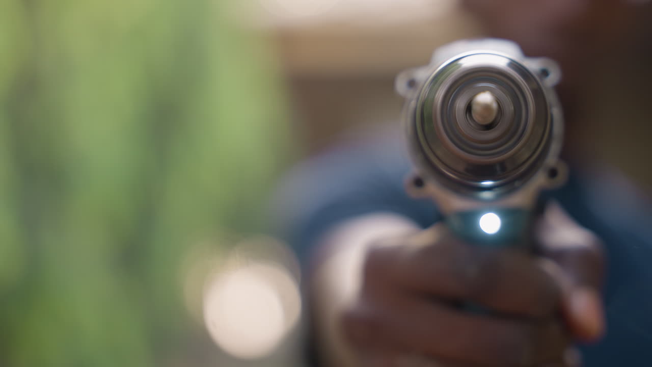 Closeup power drill aimed at camera, carpentry apprentice holds cordless drill outdoors, hand and chuck in sharp focus, blurred green background, led light glowing, workshop vibe, diy repair scene
