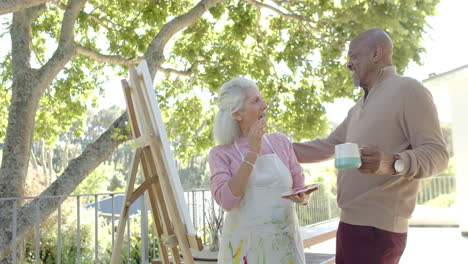 happy senior biracial couple painting and drinking coffee at balcony at home, slow motion