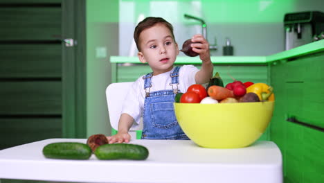 adorable toddler kid in jeans romper sits in the kitchen. toddler picks a vegetable from a bowl full of veggies and puts on the table.