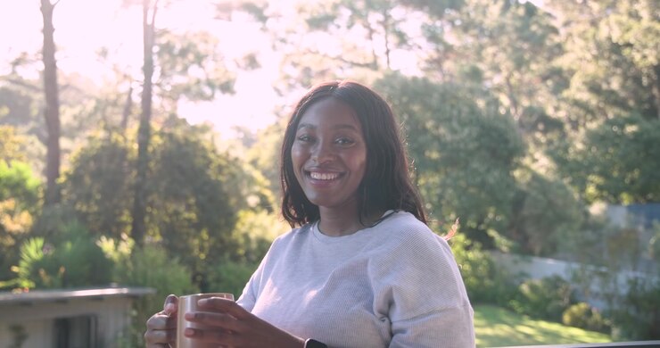 African woman leaning on balcony railing holding mug, reacting to camera tightening, smiling