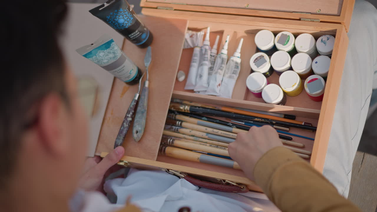 artista organizando caixa de tinta e pincéis, preparando-se para sessão de esboços de rua, mãos selecionando tubos e potes em organizador de madeira, luz solar, postura focada, roupas casuais, ambiente de varanda urbana