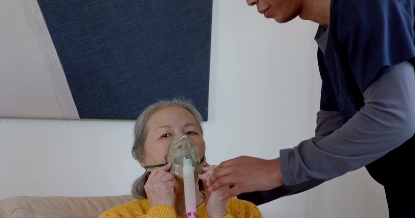 Operatore sanitario in divisa che si china, applicando il tubo della maschera per l'ossigeno a un'anziana donna cinese sul divano per aiutarla a respirare.