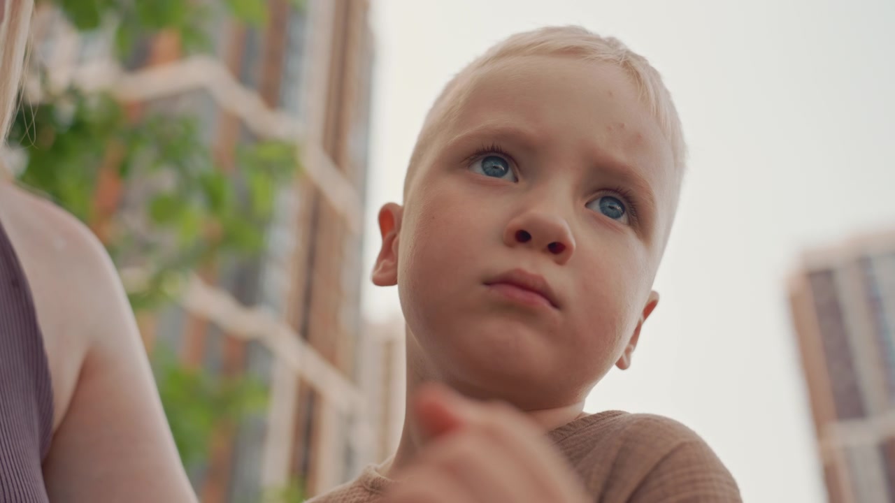 Menino jovem de cabelo claro é segurado por uma mulher que olha para a câmera. O fundo mostra um cenário urbano com edifícios e vegetação visíveis sob iluminação suave de luz do dia.