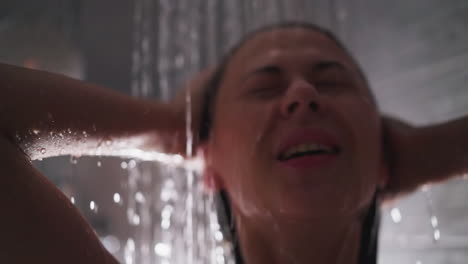 woman under clear water in shower cabin closeup. lady with closed eyes strokes wet hair under warm sprays in bathroom. spa care for rest at home