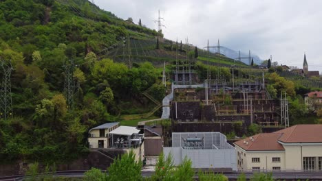 electric substation at the base of terraced vineyards in bolzano, where pylons climb the green hillsides under a cloudy sky, blending urban energy structures near via santa maddalena di sotto.