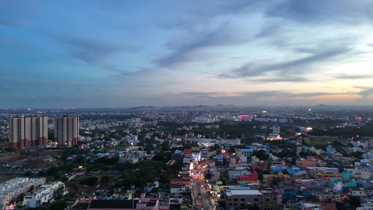 Il panorama urbano di Chennai al crepuscolo mostra le fitte strade più antiche in primo piano e i grattacieli residenziali sullo sfondo. I lampioni stradali e le scie del traffico illuminano la scena, con le colline lontane visibili all'orizzonte.