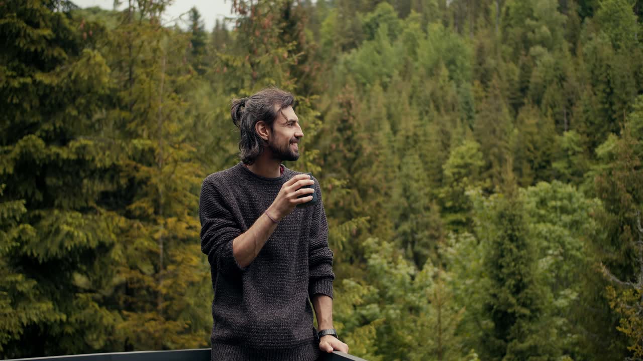 visión general de un tipo moreno feliz que está de pie en el balcón, bebiendo té y mirando el bosque de coníferas en ogros