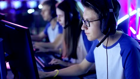 professional boy gamer plays in video game on a esports tournament/ internet cafe. he wears glasses and headphones and speaks into microphone. other girls and boys players playing in background.