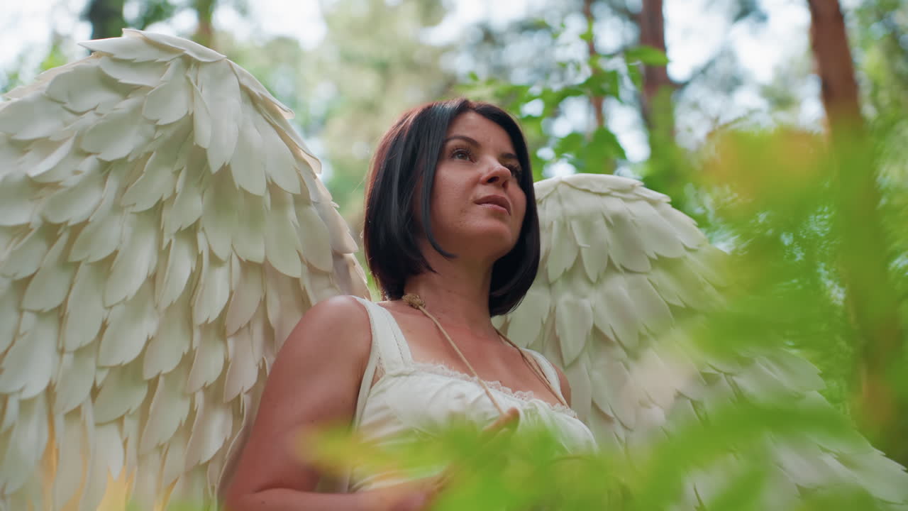 Close up of woman with large white angel wings walking through peaceful forest while gently beating hand drum, sunlight shimmering on feathers and skin, evoking calm rhythm