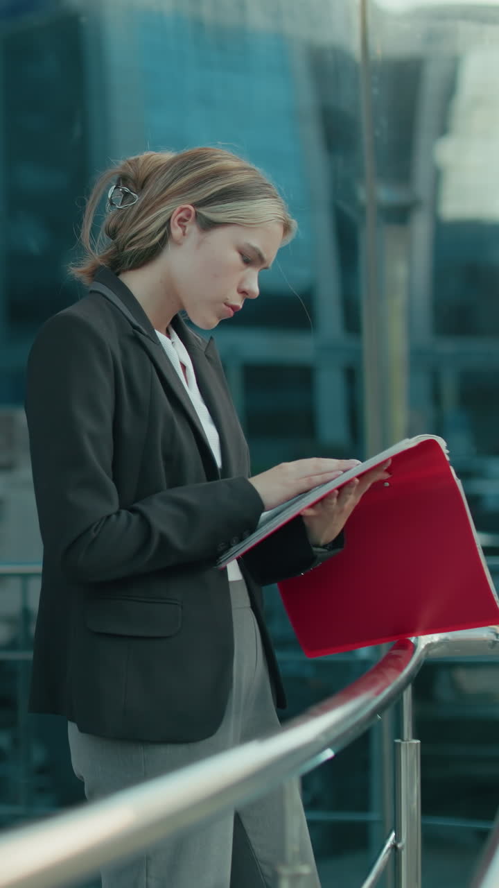 Young ceo reading red folder while standing beside iron railing in modern urban setting with glass office building reflecting surroundings including parked cars and sunlight casting bright tone