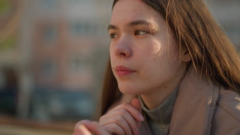 a young girl in a peach jacket sits in a park, deep in thought with her chin resting on her hand. her expression is reflective, as she meditates quietly in the peaceful outdoor setting