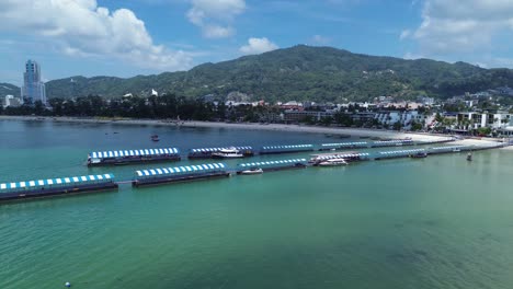 drone flies up behind a rock, unveiling the stunning patong beach with turquoise waters and swimmers enjoying the sunny coastline of phuket, thailand.