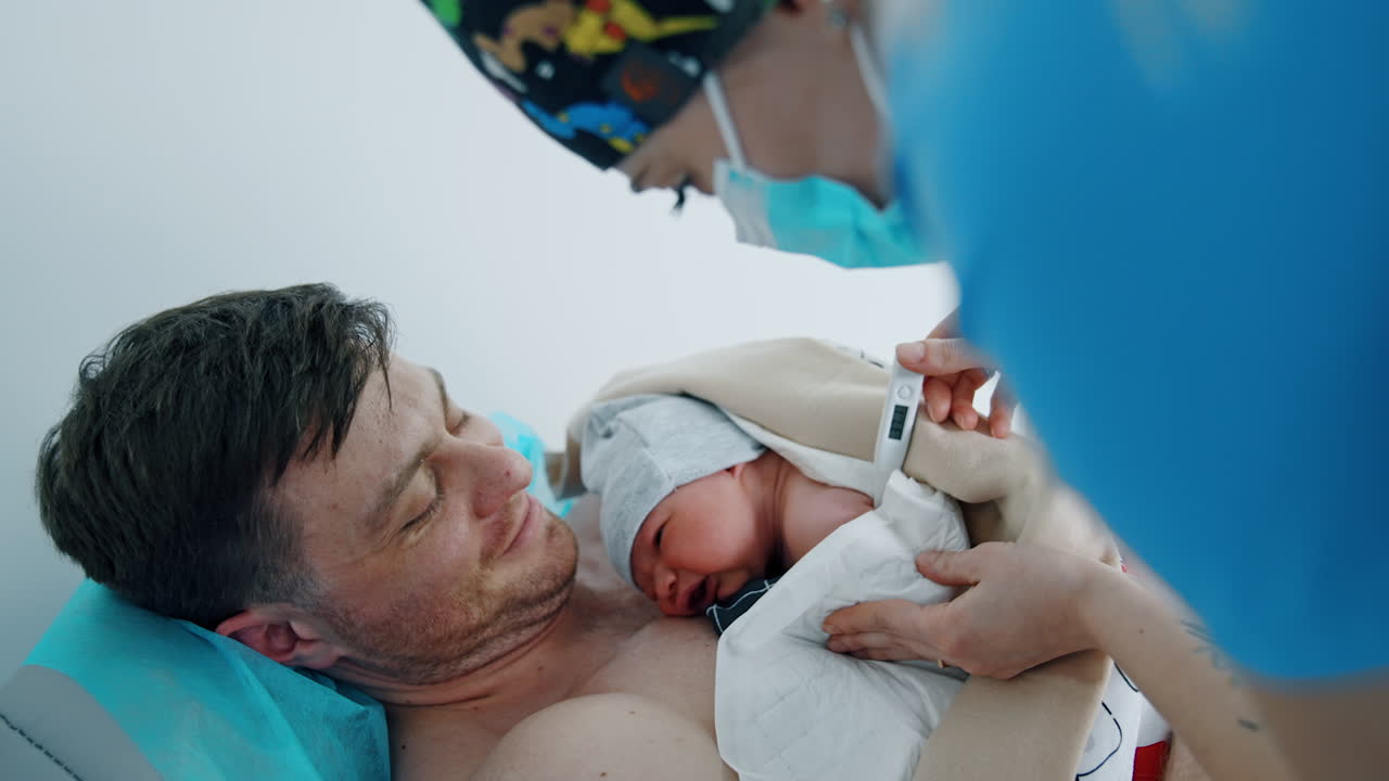 Happy father is holding his newborn child on the chest. Medical nurse is taking baby's temperature.