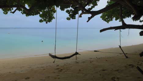 Eine-Leere-Holzschaukel-An-Einem-Tropischen-Strand,-Die-Im-Wind-Schwingt,-Mit-Blauem-Meer-Im-Hintergrund