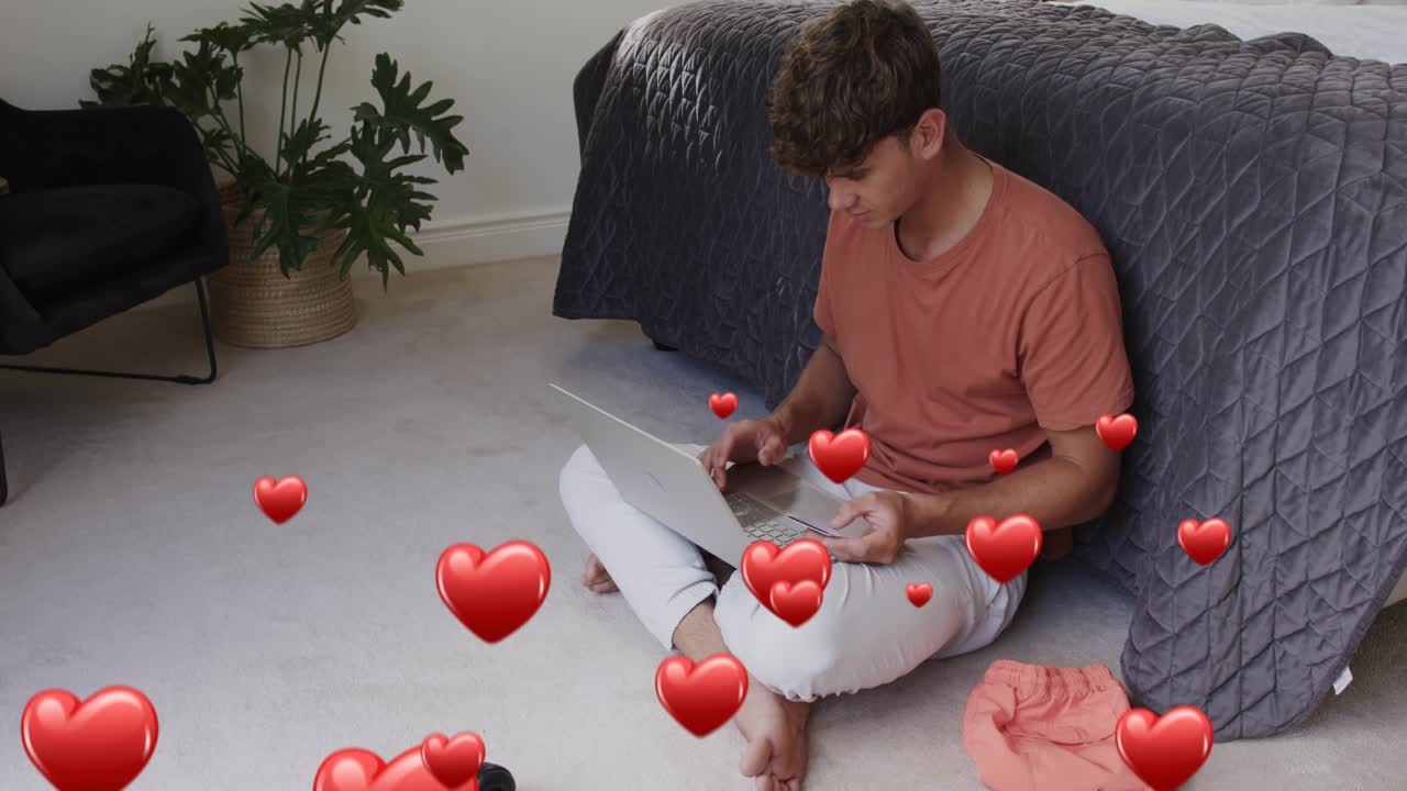 Man sitting on bedroom carpet tapping phone and typing laptop creating social app hearts around him