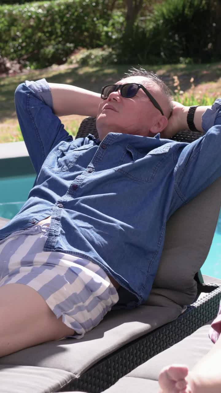 Vertical video: Stretching then resting, man leaning back on pool lounger in blue shirt, sunglasses