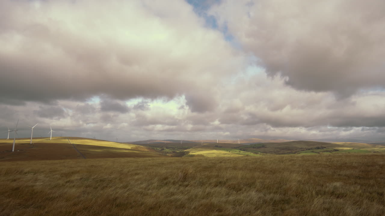Open Wide Landscape View with Wind Farm Turbines to Left and Open Farmed Welsh Countryside to the Right with Grass Blowing in Heavy Wind