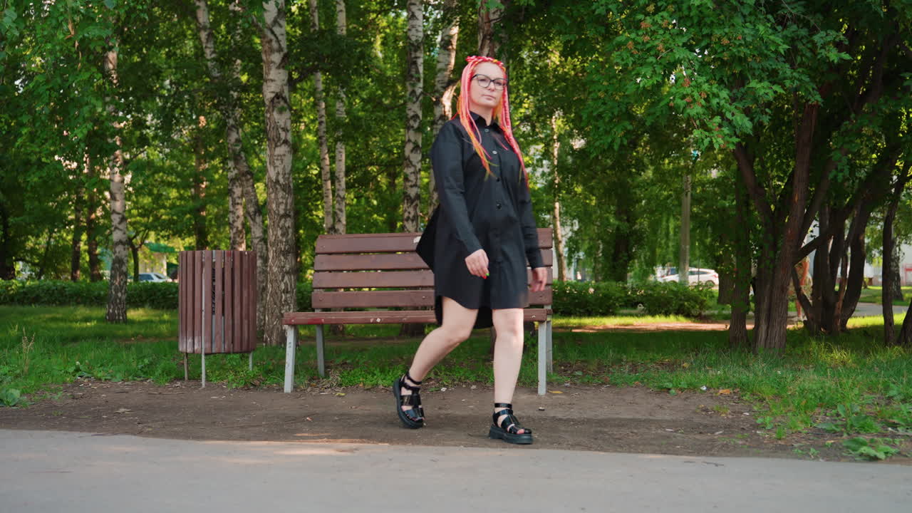 여름 옷을 입은 여성, 스타일리시한 옷차림으로 공원 벤치와 쓰레기통 근처를 걷는 빨간 머리 여성, 땋은 빨간 머리를 한 패셔너블한 여성이 여름의 푸른 녹음 속에서 공원 가구를 지나 걸어가고 있어요.