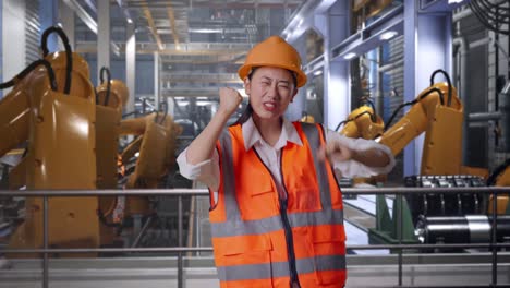 asian female engineer with safety helmet screaming goal and dancing celebrating with industrial robotic arms working