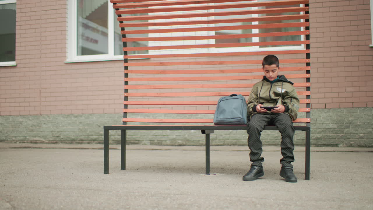 한 어린 소년이 벤치에 앉아 쉬는 시간에 휴대폰 게임을 하고 있어요. 옆에는 가방이 있고, 화면에 다시 집중하기 전에 왼쪽과 오른쪽을 흘끗 봅니다. 현대적인 벤치와 벽돌 벽이 있는 고요한 야외 학교 운동장 풍경이에요.