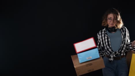 hermosa mujer sonriente con camisa a cuadros y vaqueros caminando alegremente con coloridas bolsas de compras, posando juguetón sobre un fondo negro. con gafas y auriculares negros en el cuello. cámara lenta