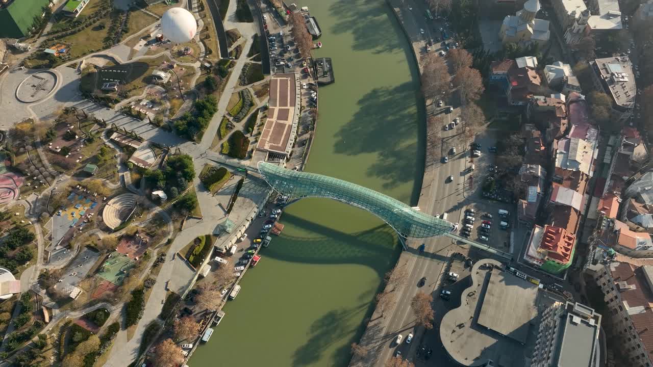 Aerial view featuring Rike Park, the Bridge of Peace, and green riverbanks in central Tbilisi. The drone shot highlights the urban design, walking areas, and mix of nature with architecture