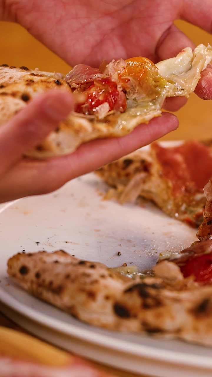 Mani sollevano una fetta di pizza fresca con prosciutto e pomodorini da un piatto bianco.